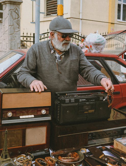 An elderly man with a white beard, wearing a grey cap, sunglasses, and a grey zip-up sweater over a checked shirt, is standing next to a red car on a city street. He is holding a beaded bracelet in hi