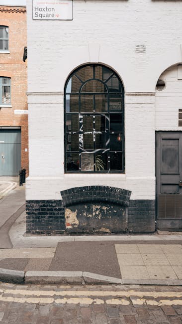 The image shows the exterior of a white-painted brick building with an arched, black-framed window occupying most of the visible wall space. Below the window, there is a black brick section that appea